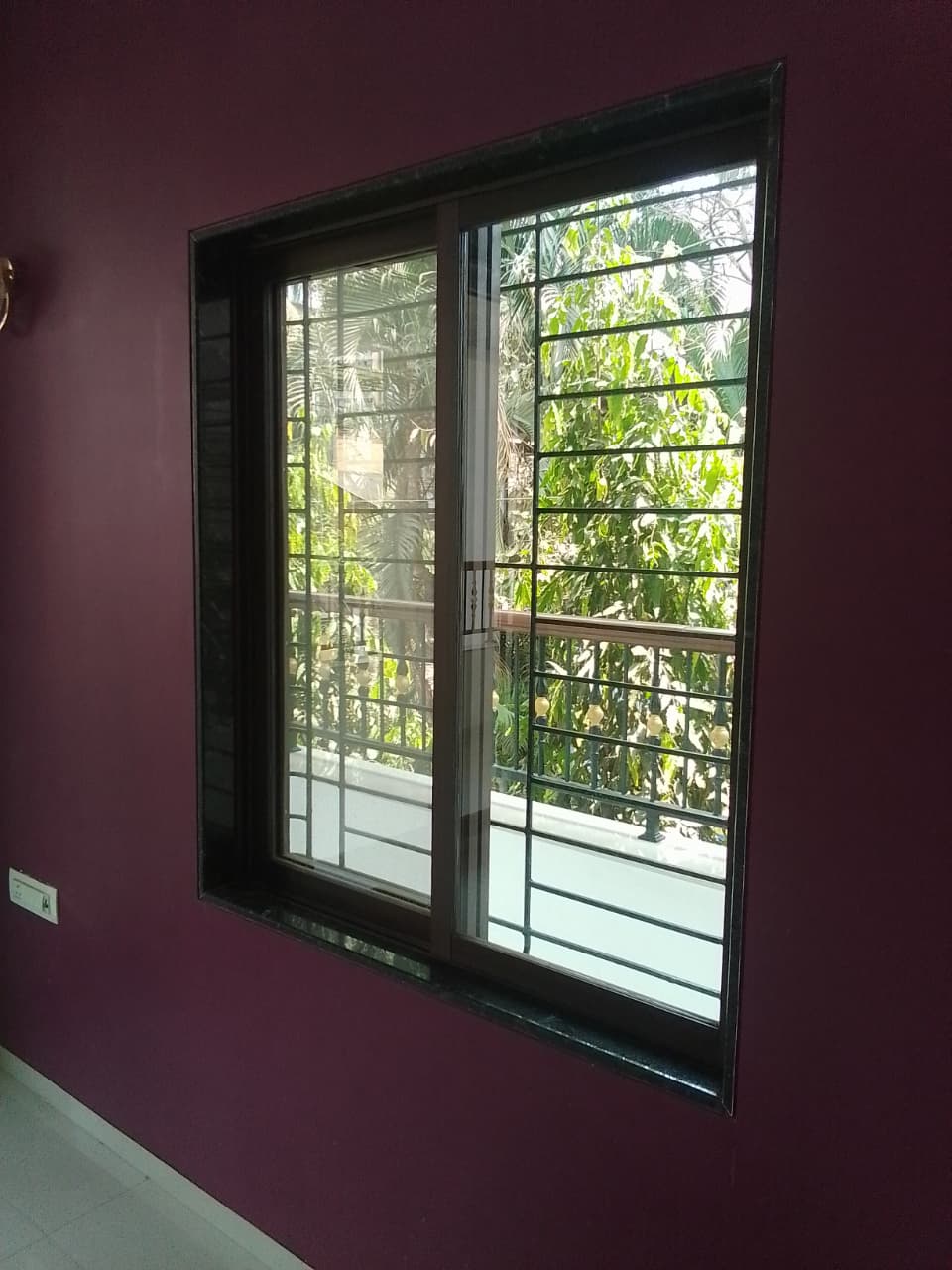 Untreated sliding window with safety grille in a purple-walled room, looking onto neighbouring foliage and a walkway