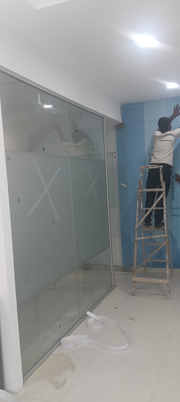 Half-height frosted film on a glass cabin partition while a worker on a ladder applies a blue printed wall graphic behind