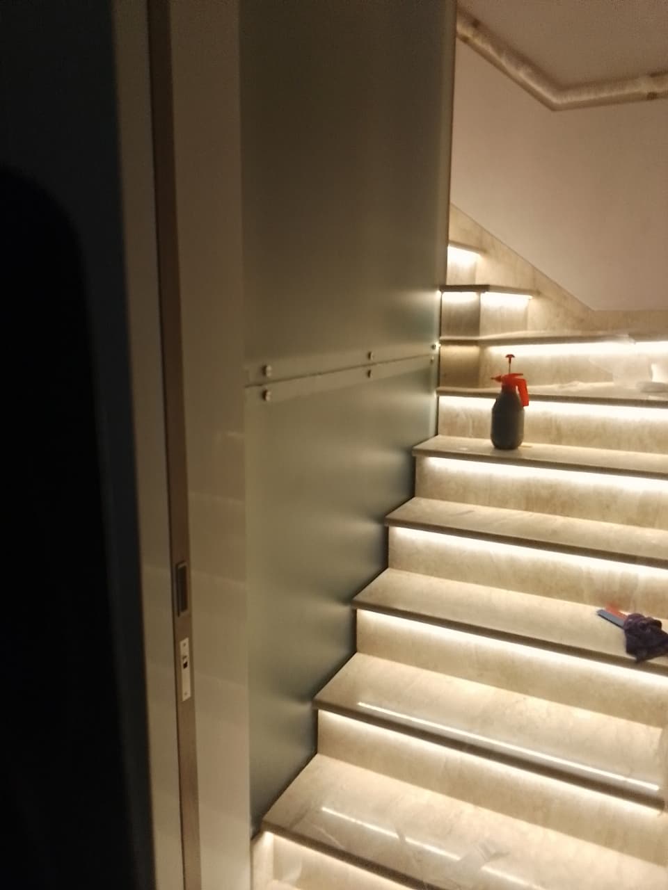 Frosted glass balustrade beside an LED-lit marble staircase, spray bottle resting on a tread during installation