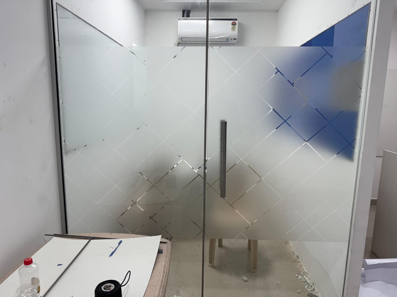 Diamond lattice frosted film being fitted to a glass partition and door, cutting table and tape roll in the foreground