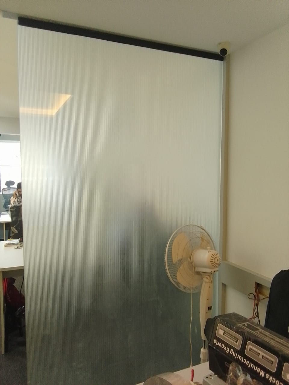 Full-height reeded translucent film on an office partition screening a workstation area, wall fan and desks beyond