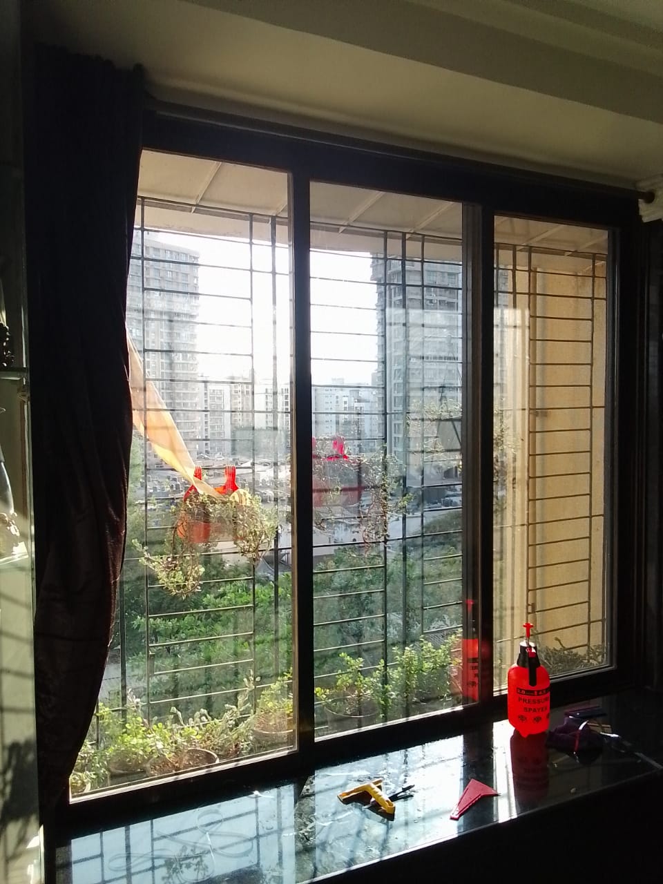 Three-track aluminium sliding windows in a Mumbai flat during window film application, with squeegee, blade and spray bottle on the sill