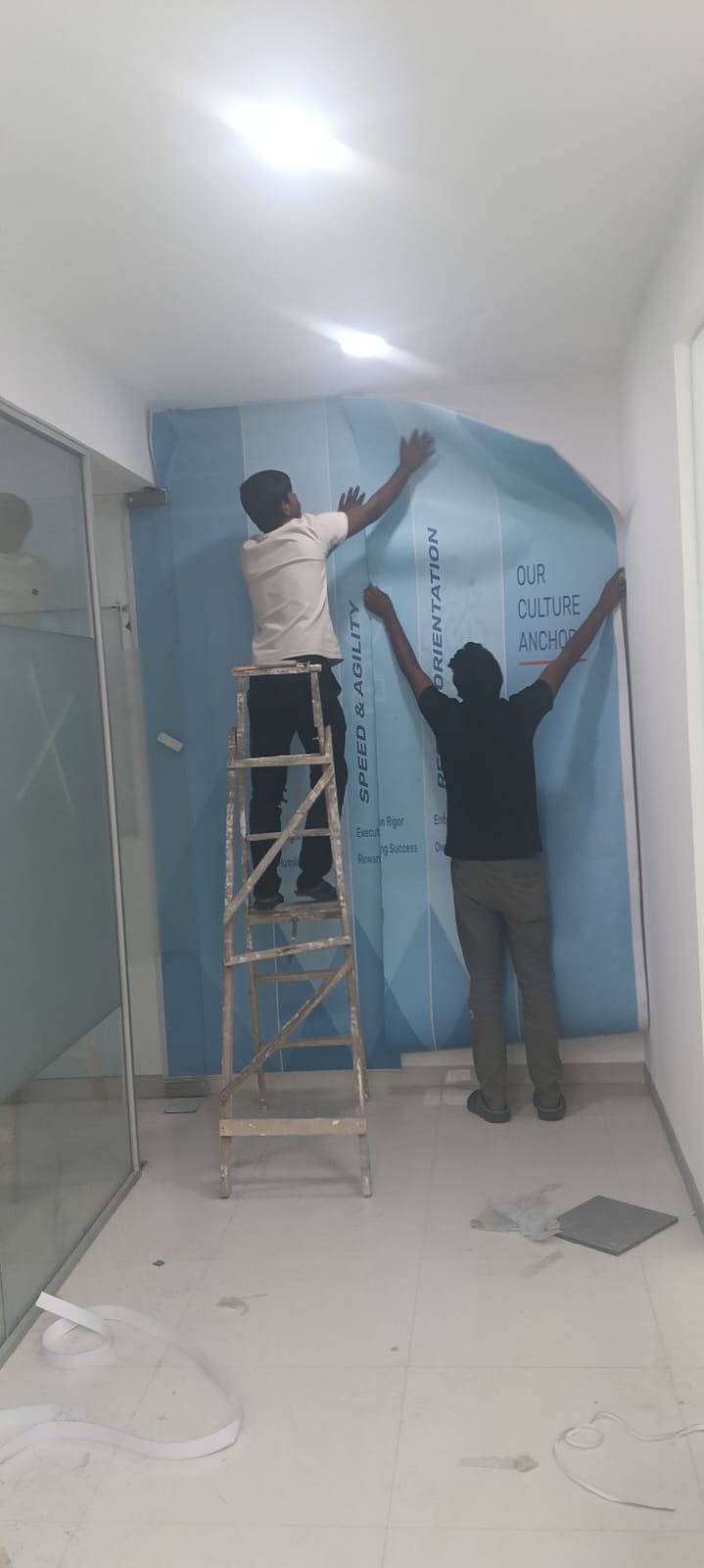 Two installers smoothing a large printed corporate culture wall graphic onto an office wall from a ladder